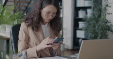 Genç Asyalı iş kadını akıllı telefon mesajı kullanıyor. İnternette çalışıyor. Ofiste oturuyor. Modern alet ve profesyonel meslek kavramı.