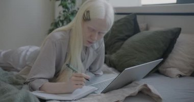 Yatak odasında yatan dizüstü bilgisayarla öğrenen albino kız öğrencilerin yavaş çekimleri. İnternet eğitimi ve gençlik kavramı.