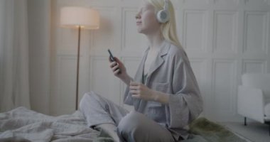 Neşeli albino kadın, kulaklıkla müzik dinleyip rahat bir dairede yatağında akıllı telefon tutuyor. Modern yaşam tarzı ve eğlence konsepti.