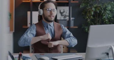 Yaratıcı genç iş adamı kulaklıkla müzik dinliyor ve ofiste oturmuş dans ediyor. İş yeri ve eğlence konsepti.