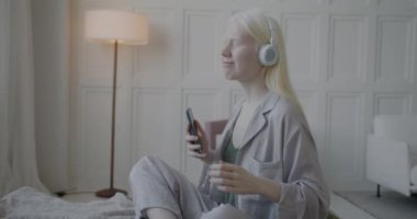 Pijamalı mutlu albino kızın kulaklıkla müzik dinlemesi ve lüks yatak odasında yatakta akıllı telefon tutarak dans etmesi. Gençlik kültürü ve eğlence konsepti.