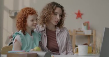 Anne ve kızı dizüstü bilgisayarla çevrimiçi video görüşmesi yapıyor. Evde el sallayarak sohbet ediyorlar. Sanal toplantı ve aile kavramı.