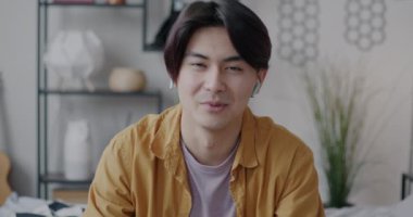 Evdeki yatak odasında kablosuz kulaklık kullanarak kameraya bakarak internetten konuşan neşeli Asyalı adamın portresi. İnsanlar ve iletişim konsepti.