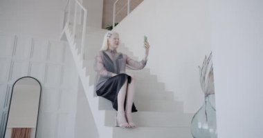 Çekici albino kadın, lüks evdeki merdivenlerde otururken çevrimiçi video görüşmesi yapıyor. Mesafe iletişimi ve şık iç mekan kavramı.