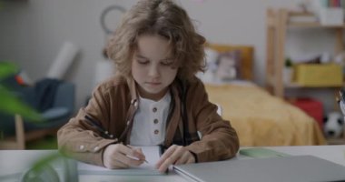 Masasında kalem ve defterle oturan sevimli çocuğun yavaş çekimde portresi. Sonra da yatak odasında kameraya bakıp gülümsüyor. Çocukluk ve ev kavramı.