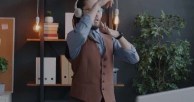 İş yerinde dans eden, kulaklıkla kameraya bakan çekici genç ofis çalışanının yavaş çekim portresi. İş ve eğlence konsepti.