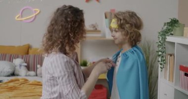 Evde maskeli balo partisi için oğlunu hazırlayan küçük çocuğa süper kahraman kostümü giydiren genç bir kadın. Çocukluk ve eğlence kavramı.