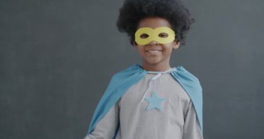 Süper kahraman kostümlü mutlu Afro-Amerikan çocuk portresi gri arka plandaki kameraya gülümseyen el hareketlerini gösteriyor. Olumlu duygu ve çocukluk kavramı.