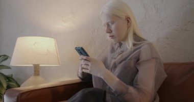 Zeki telefonlu çekici albino kız, elinde modern bir aletle evde şık bir odada deri koltukta oturuyor. İletişim ve insan kavramı.