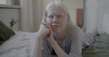 Yavaş çekimde, pijamalı, kalp hareketi yapan ve lüks yatak odasında yatağında sırıtan sevimli albino kadının portresi. Aşk ve şefkat kavramı.