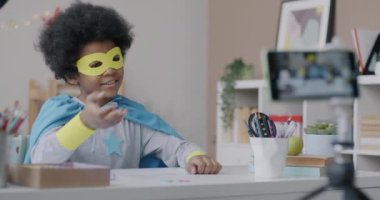 Süper kahraman maskeli ve pelerinli komik Afrikalı çocuk evdeki akıllı telefon kamerasını kullanarak internet bloğu için video içeriği kaydediyor. Günlükler ve çocukluk kavramı.