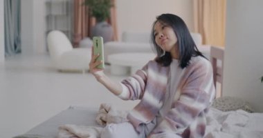 Akıllı telefon kamerası için poz veren çekici Asyalı kadının lüks bir dairede yatakta selfie çekmesi. Sosyal medya ve fotoğrafçılık kavramı.
