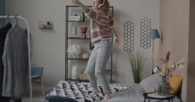 Yatakta dans eden neşeli genç bir kadın kulaklıkla müzik dinliyor yatak odasında eğleniyor. Modern yaşam tarzı ve eğlence konsepti.