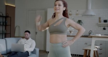 Genç kadın evde spor yaparken koca dizüstü bilgisayarla çalışıp evdeki kanepede oturan karısına bakıyor. Etkin yaşam tarzı ve ilişki kavramı.