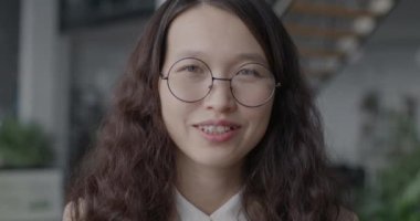 Ofisteki kameraya gülümseyen neşeli Asyalı genç iş kadınının yavaş çekim portresi. İş ve insan kavramı.