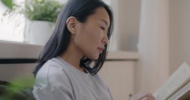 Mutlu Asyalı kadın kitap okurken yavaş çekimde gülümsüyor evdeki şık, aydınlık yatak odasında rahat bir yatakta oturuyor. Edebiyat ve eğlence konsepti.