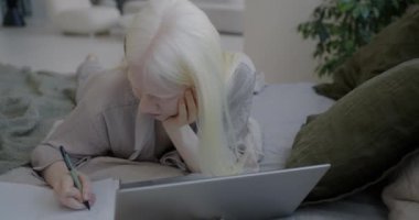 Öğrenci albino kızın dizüstü bilgisayarla bilgi araması ve not defterine yazması yatak odasındaki sanal eğitime odaklıydı. Mesafe öğrenme ve gençlik kavramı.