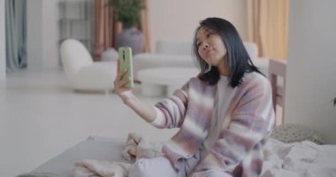 Akıllı telefon kamerası için poz veren çekici Asyalı kadının lüks bir dairede yatakta selfie çekmesi. Sosyal medya ve fotoğrafçılık kavramı.