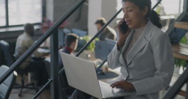 Orta Doğulu iş kadını cep telefonuyla konuşuyor ve açık uzay ofisindeki merdivenlerde çalışırken dizüstü bilgisayar kullanıyor. İş adamı ve iletişim konsepti.