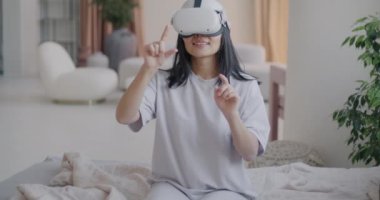 Sanal gerçeklik gözlüğü takan genç bir bayanın lüks bir dairede yatakta otururken el kol hareketi yapması. Modern teknoloji ve yaşam tarzı kavramı.