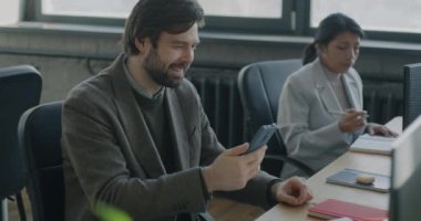 Sanal toplantı sırasında iş ortağıyla konuşan neşeli genç bir işadamı ofisteki akıllı telefondan online video görüşmesi yapıyor. İnternet iletişimi ve iş kavramı.