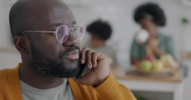 Evdeki mutfakta cep telefonuyla konuşan Afro-Amerikalı bir adam. Karısı ve oğlu arka planda görünüyor. İletişim ve aile yaşam tarzı kavramı.