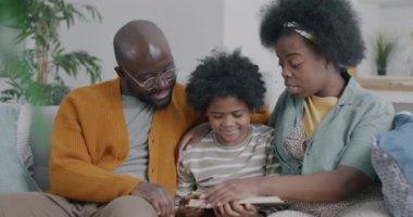 Genç Afrikalı Amerikalı kadın, çocuk ve kocasıyla kanepede oturan küçük oğluna kitap okuyor. Edebiyat ve aile yaşam tarzı kavramı.