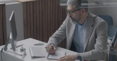 Deneyimli bir iş adamı bilgisayar ekranında araştırma yapıyor ve ofiste not defteri yazıyor. İş ve iş kavramı.