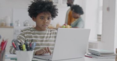 Ebeveynler arka planda mutfakta yürürken, evde çalışan Afrikalı Amerikalı çocuk dizüstü bilgisayarla daktilo kullanıyor. Evde eğitim ve eğitim kavramı.