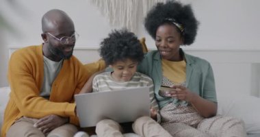 Neşeli Afro-Amerikan aile laptopları ve kredi kartlarıyla alışveriş yapıyor. Evde yatakta oturmuş gülüyorlar. Tüketim ve e-ticaret kavramı.