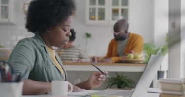 Afrikalı Amerikalı kadın dizüstü bilgisayar kullanıyor ve not defterine bilgi yazıyor. Bu arada arka planda oğluyla meşgul bir adam var. İnternet bağlantısı ve serbest meslek kavramı.