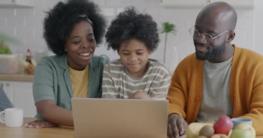 Anne baba ve çocuk dizüstü bilgisayar kullanarak konuşuyor ve gülüyorlar. Birlikte mutfakta otururken ekrana bakıyorlar. Aile yaşam tarzı ve modern teknoloji kavramı.