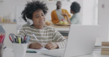 Afro-Amerikalı öğrenci sanal sınıfta öğreniyor. Ebeveynler mutfakta konuşurken kulaklık ve dizüstü bilgisayarla öğretmenleri dinliyor. Eğitim ve insanlar kavramı.