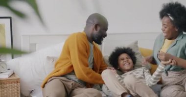 Afrikalı Amerikalıların ebeveynleri küçük çocuğu gıdıklayıp gülüyor sonra da evde yatakta yatan çocuğa sarılıyorlar. Aile eğlencesi ve duygu konsepti.