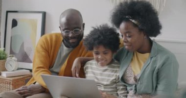 Neşeli Afro-Amerikan ailesi dizüstü bilgisayar kullanarak ekranda konuşarak ve gülerek evdeki yatakta oturarak internet içeriğini tartışıyor. İnsanlar ve teknoloji kavramı.