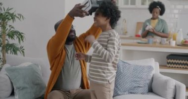 Küçük çocuk babasıyla sanal gerçeklik gözlüğü kullanırken annesi evde mutfakla meşguldü. Aile yaşam tarzı ve modern teknoloji kavramı.