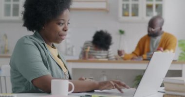 Genç Afro-Amerikalı kadın dizüstü bilgisayarı ve kredi kartıyla alışveriş yaparken kocası ve çocuğu mutfakta görünüyor. Tüketim ve aile yaşam tarzı kavramı.