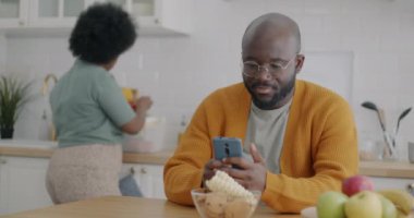 Genç Afro-Amerikalı adam akıllı telefon kullanırken karısı mutfakta yemek pişiriyor. Modern teknoloji ve aile yaşam tarzı kavramı.