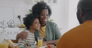 Anne, oğluna sarılıp, babasıyla konuşuyor ve mutfakta aile kahvaltısı yapıyor. Annelik ve ilişki kavramı.