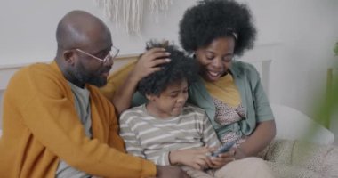 Mutlu Afro-Amerikan ailesi akıllı telefon kullanarak evde oturup gülüyor ve konuşuyor. Modern teknoloji ve insan kavramı.