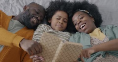 Neşeli Afrika kökenli Amerikalı aile babası ve çocuğu kitap okuyor ve evde yatıp gülüyor. Edebiyat ve eğlence konsepti.