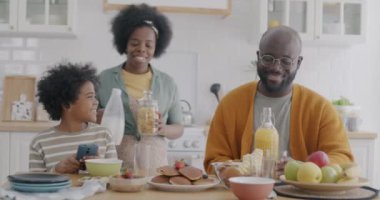 Genç Afro-Amerikan kadın kahvaltı için yemek getiriyor ve kocasıyla konuşurken oğlu da mutfakta akıllı telefon kullanıyor. Aile yaşam tarzı ve beslenme konsepti.