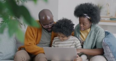 Mutlu Afrikalı Amerikalı çocuk dizüstü bilgisayar kullanıyor ve anne ve babasıyla konuşuyor. Evde kanepede oturuyor. Teknoloji ve çocukluk kavramı.