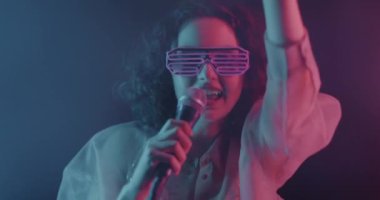 Komik gözlüklü bir kadının portresi. Mikrofonda şarkı söylüyor. Karanlık arka planda neon ışıkta dans ediyor. Eğlence ve gece hayatı konsepti.