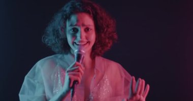 Mikrofonda şarkı söyleyen kadının karanlık arka planda neon ışıkta dans eden kamerayı göstermesi. İnsanlar ve eğlence konsepti.