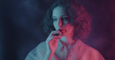 Elektronik sigara içen genç bir kadın karanlık arka planda neon ışıkta dinleniyor. Buhar makinesi ve modern yaşam tarzı konsepti.