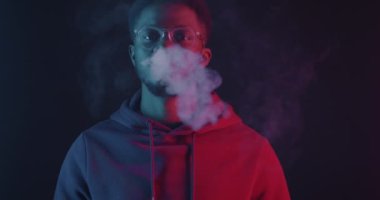 Siyah arka planda, neon ışıkta kameraya bakan Afro-Amerikalı bir adam. Modern sigara aleti ve elektronik sigara konsepti.