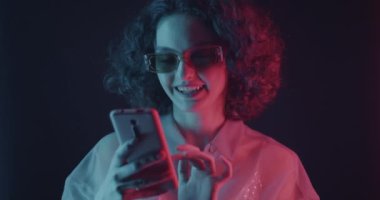 Akıllı telefon uygulaması kullanan genç bir kadın karanlık arka planda neon ışıkta gülümsüyor. Modern teknoloji ve insan kavramı.