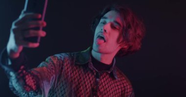 Akıllı telefon kamerası için selfie çeken genç adam karanlık arka planda neon ışıkta eğleniyor. Fotoğraf ve gece hayatı kavramı.