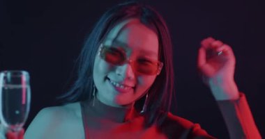 Güzel Asyalı kadın portresi kadeh kaldırarak dans ediyor ve neon ışıkta kameraya bakıyor. Eğlence ve gece hayatı konsepti.